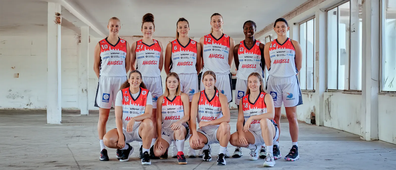 Un gruppo di otto donne in maglie da basket arancioni e bianche con la scritta 'ANGELS' posano in un magazzino.