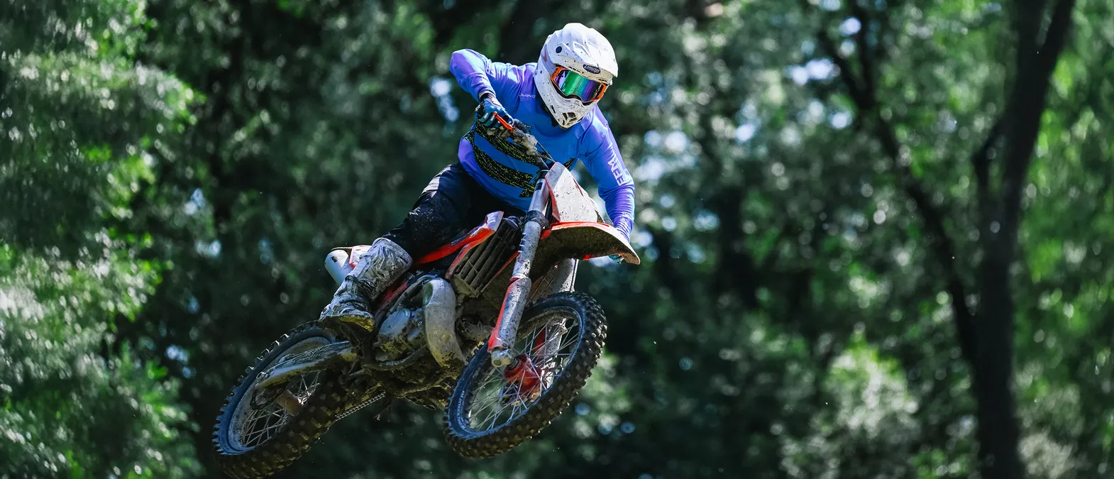 Un motociclista in tuta da corsa blu a maniche lunghe e casco bianco salta in moto attraverso una foresta.