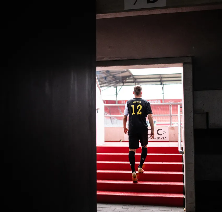 Un calciatore in una maglia nera con la scritta 'MIEYER 12' in giallo sale le scale rosse di uno stadio.