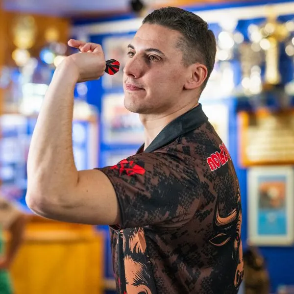 Un uomo in una maglia sportiva a motivi con dettagli rossi tiene un dardo e mira, con trofei visibili sullo sfondo.