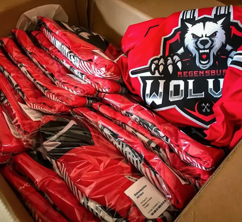 Una scatola di cartone contenente diverse maglie sportive rosse personalizzate avvolte in plastica, una delle quali con un logo di lupo e la scritta 'REGENSBURG WOLVES'.