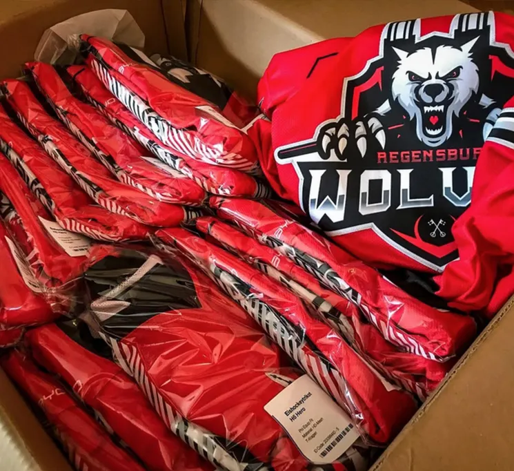 Una scatola di cartone contenente diverse maglie sportive rosse personalizzate avvolte in plastica, una delle quali con un logo di lupo e la scritta 'REGENSBURG WOLVES'.