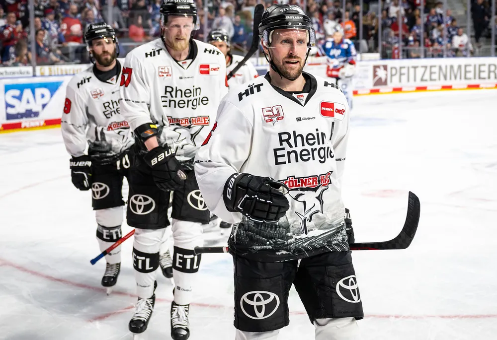 Giocatori di hockey su ghiaccio in maglie bianche con pantaloni neri che presentano il logo di una squadra, in piedi sul ghiaccio.