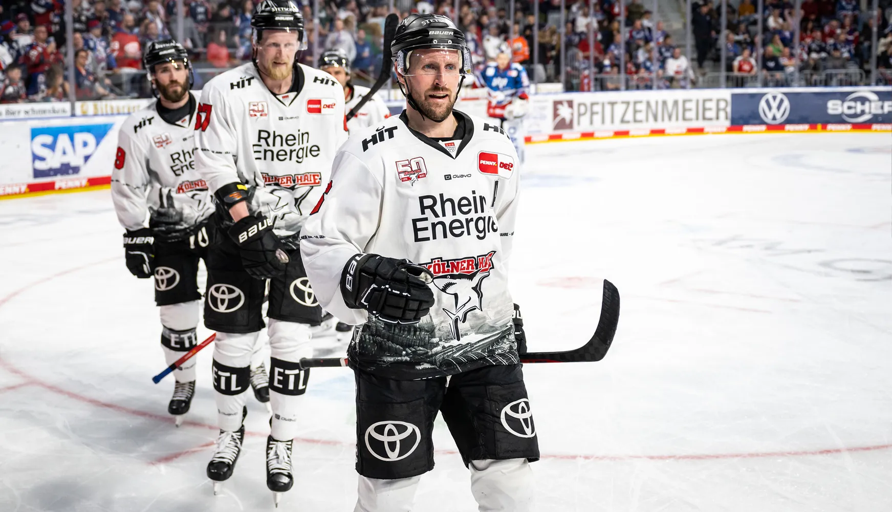 Tre giocatori di hockey su ghiaccio in maglie bianche con numeri diversi sono in piedi sul ghiaccio durante una partita.