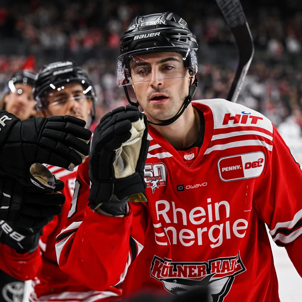 Un giocatore di hockey in una maglia rossa con strisce bianche e loghi, che alza la mano in segno di celebrazione.