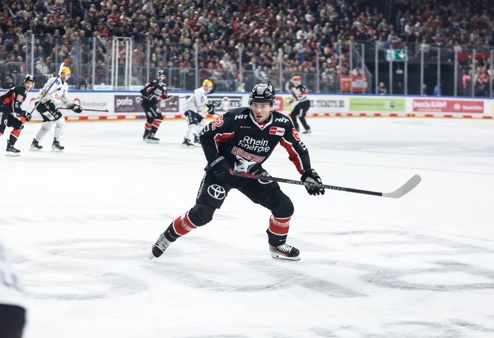 Un giocatore di hockey su ghiaccio che indossa una maglia nera con accenti rossi e bianchi, che pattina sul ghiaccio con un bastone.