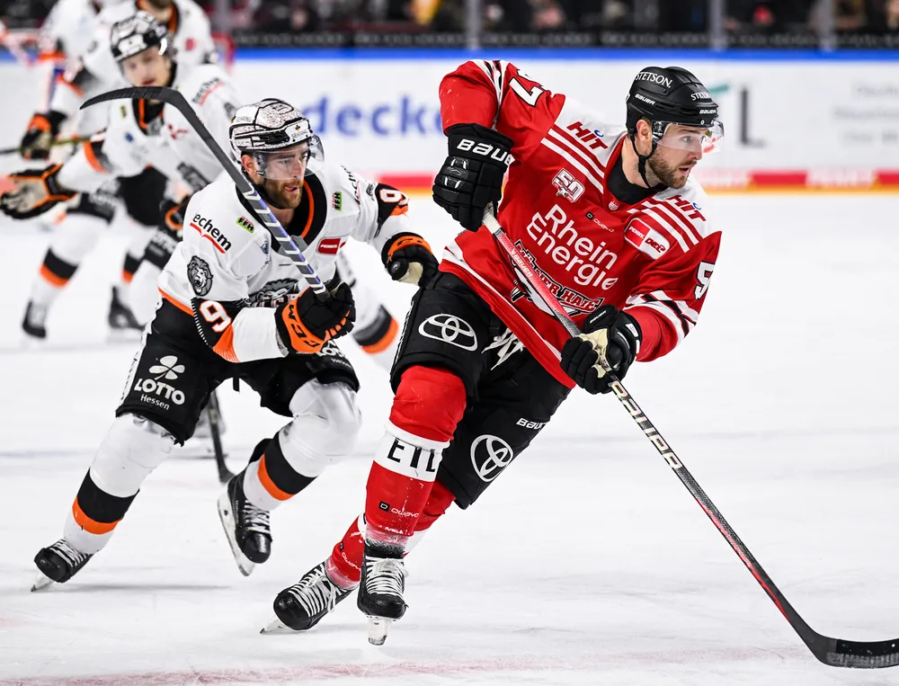 Un giocatore di hockey su ghiaccio in maglia rossa e pantaloni neri scia sul ghiaccio mentre viene inseguito da un altro giocatore.