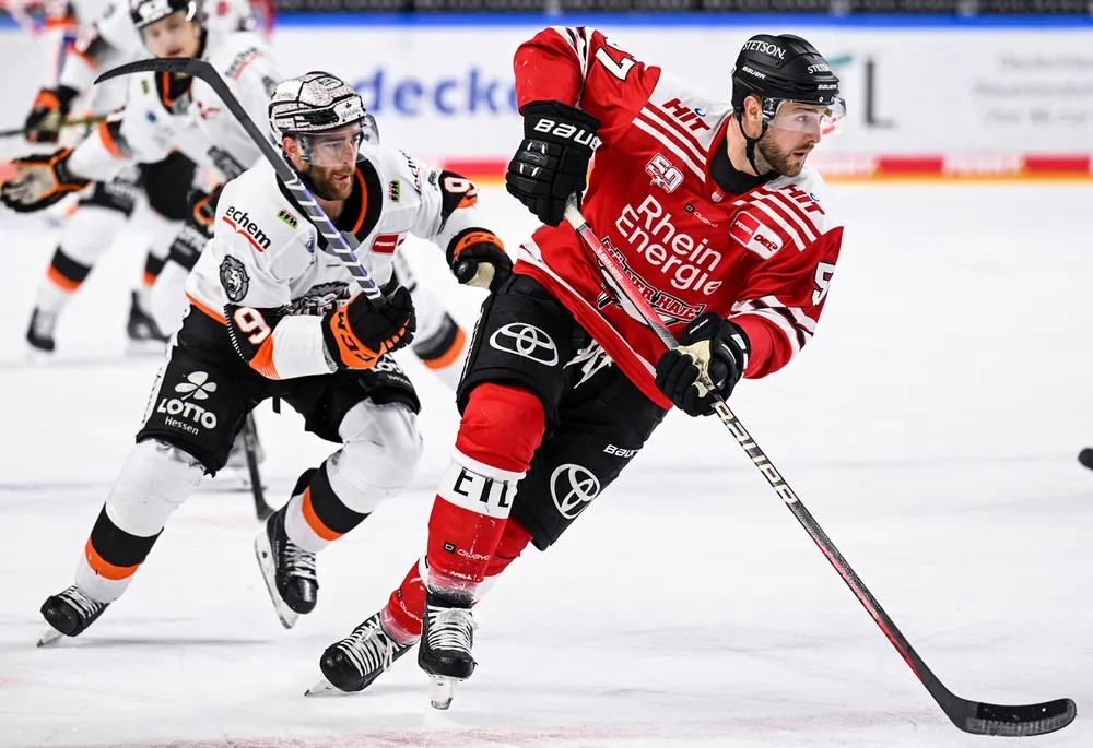 Un giocatore di hockey con una maglia rossa pattina con il puck sul ghiaccio mentre un altro giocatore con una maglia bianca lo segue da vicino.
