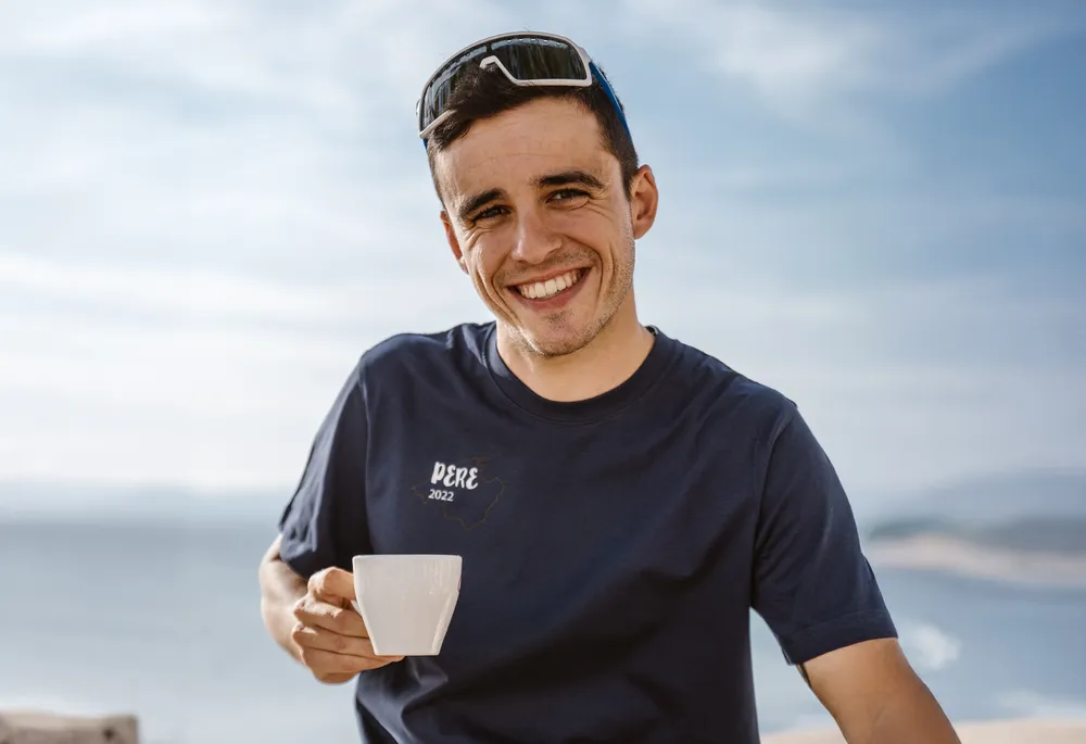 Un uomo sorridente in una maglietta blu tiene una tazza in mano, sullo sfondo di un bellissimo paesaggio costiero.