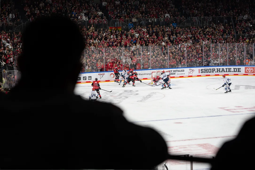 Dal punto di vista di uno spettatore, l'immagine mostra una partita di hockey su ghiaccio con giocatori in maglie rosse e blu di fronte a una grande folla.