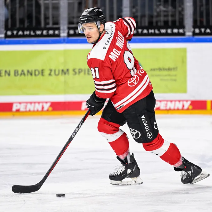 Un giocatore di hockey su ghiaccio con una maglia rossa e nera con il numero 91 e il nome Mc. Miller sul retro, in piedi sul ghiaccio e controllando un disco con la sua stecca.