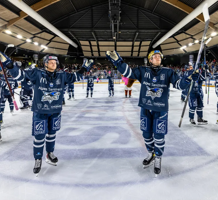 Giocatori di hockey su ghiaccio in maglie blu con loghi di sponsor festeggiano sul ghiaccio, con il pubblico sullo sfondo.