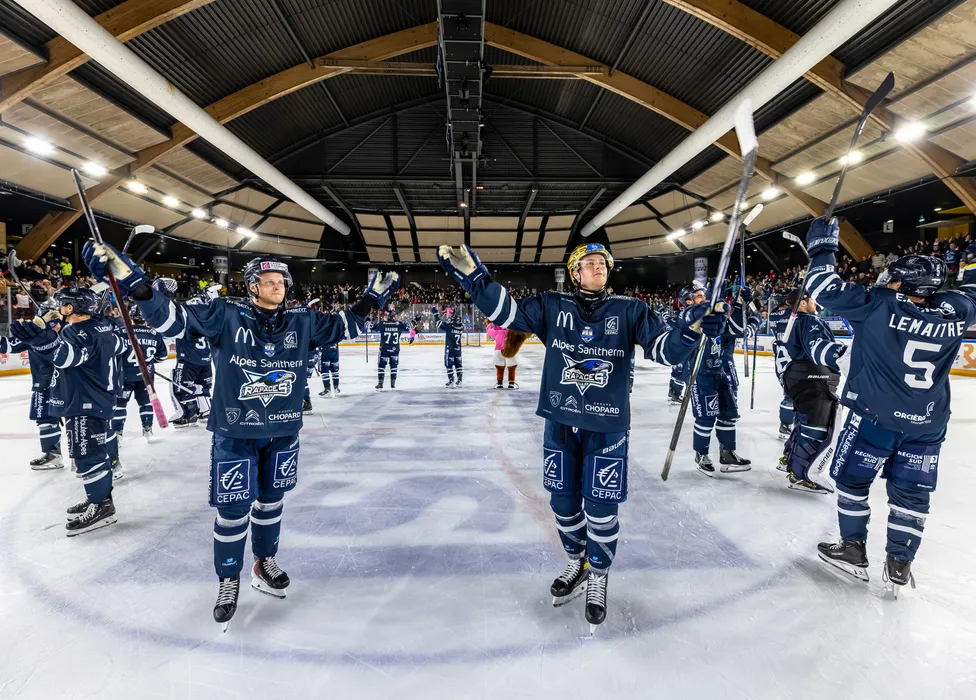 Giocatori di hockey su ghiaccio in magliette blu sono sul ghiaccio, festeggiando con i bastoni alzati di fronte a un pubblico in festa.