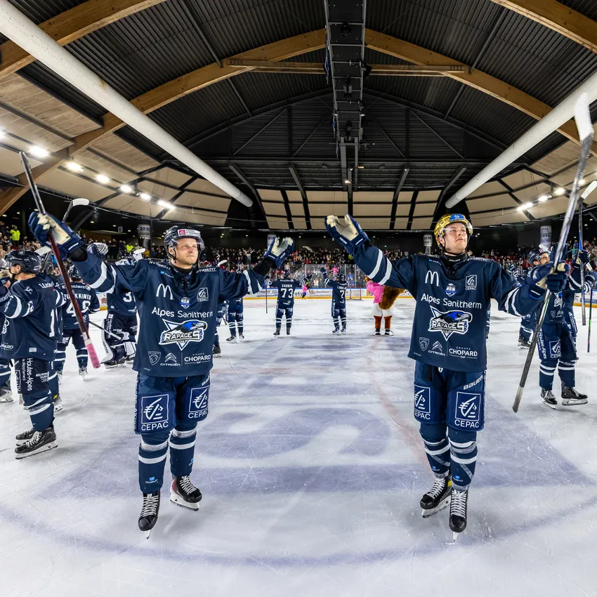 Due giocatori di hockey su ghiaccio in maglie blu in piedi sul ghiaccio, alzando le loro stecche mentre una folla in festa è visibile sullo sfondo.