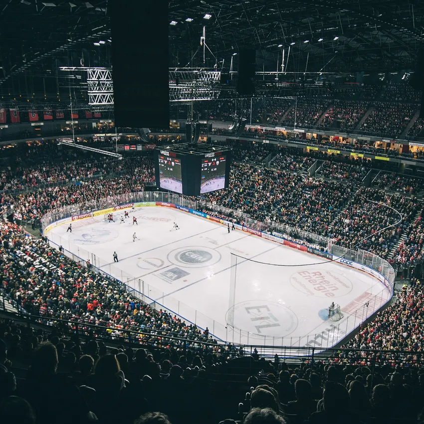 Un'arena di hockey su ghiaccio piena di tifosi che applaudono, con un campo visibile e un grande schermo sullo sfondo.