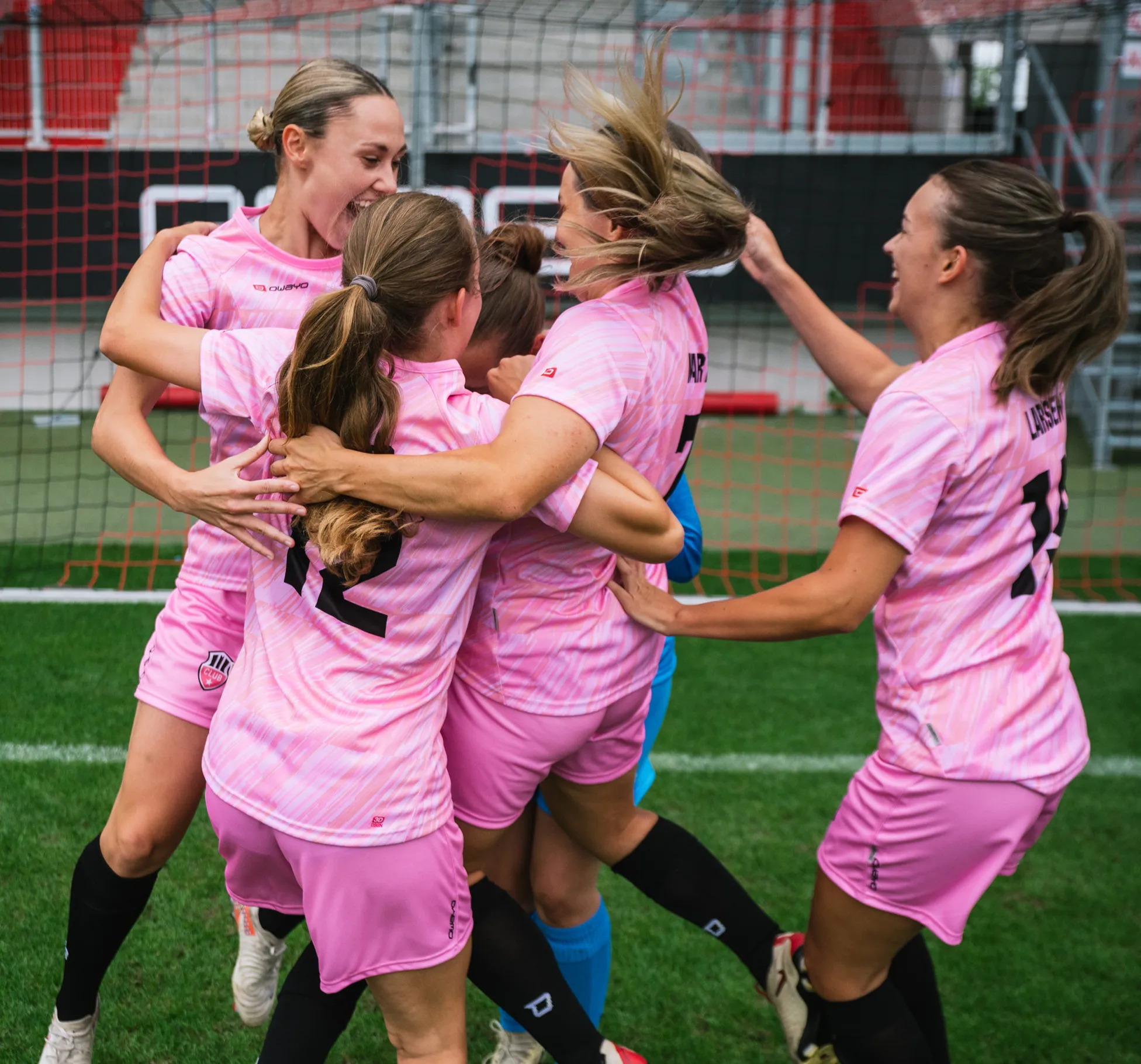 Cinque giocatrici in maglie rosa festeggiano insieme con gioia sul campo da calcio.