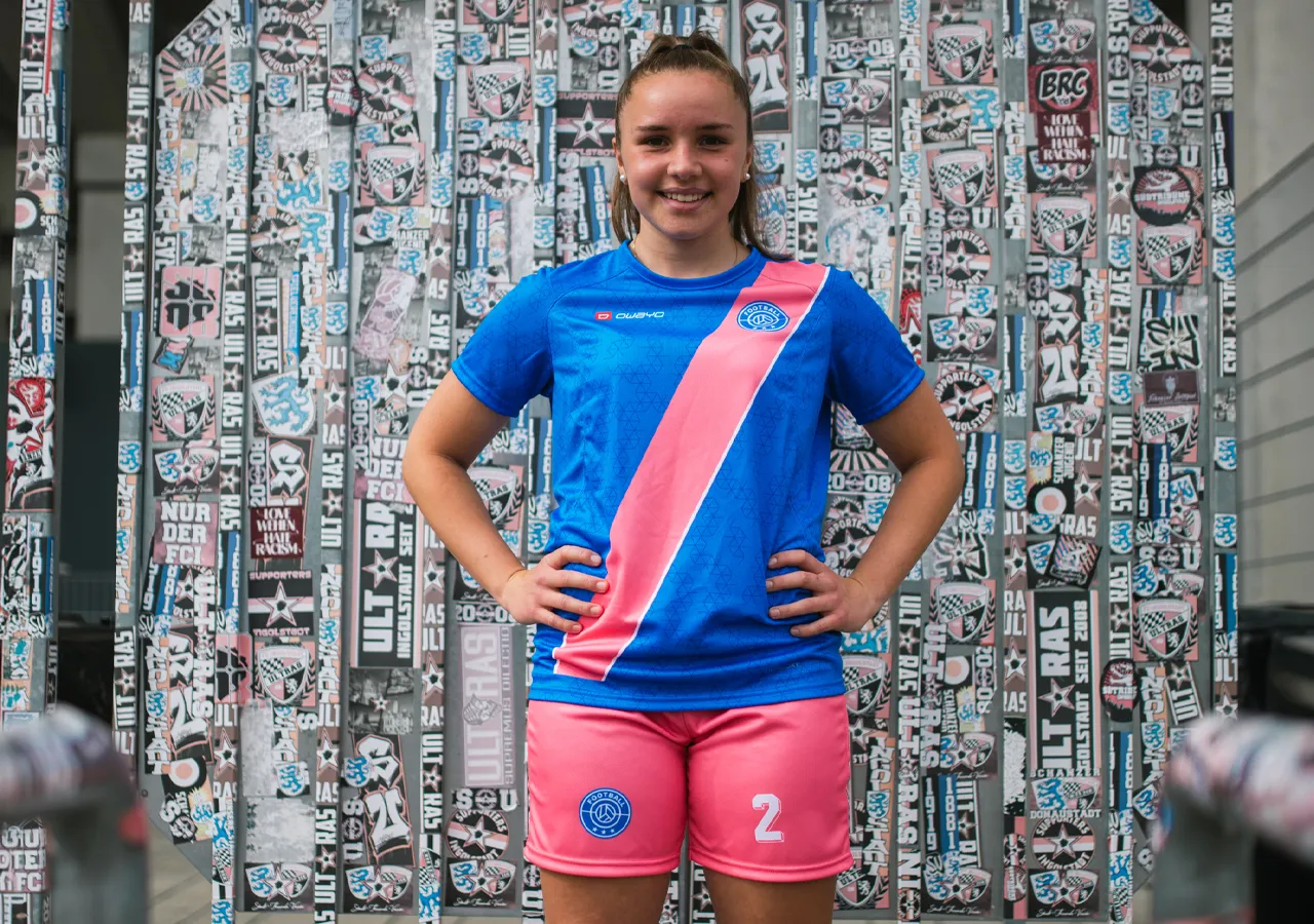 Una ragazza che indossa una maglietta da calcio blu con una striscia diagonale rosa e pantaloni corti rosa, sorridendo davanti a un muro decorato con adesivi.