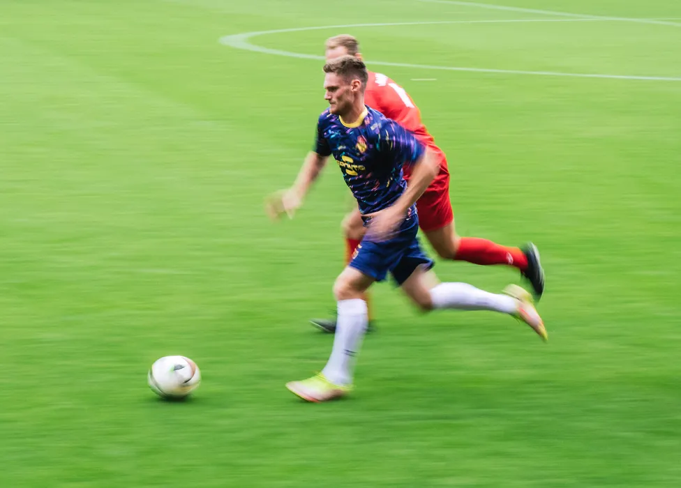 Un calciatore in una maglietta blu con motivi colorati corre rapidamente sul campo verde mentre un altro giocatore lo segue da vicino.
