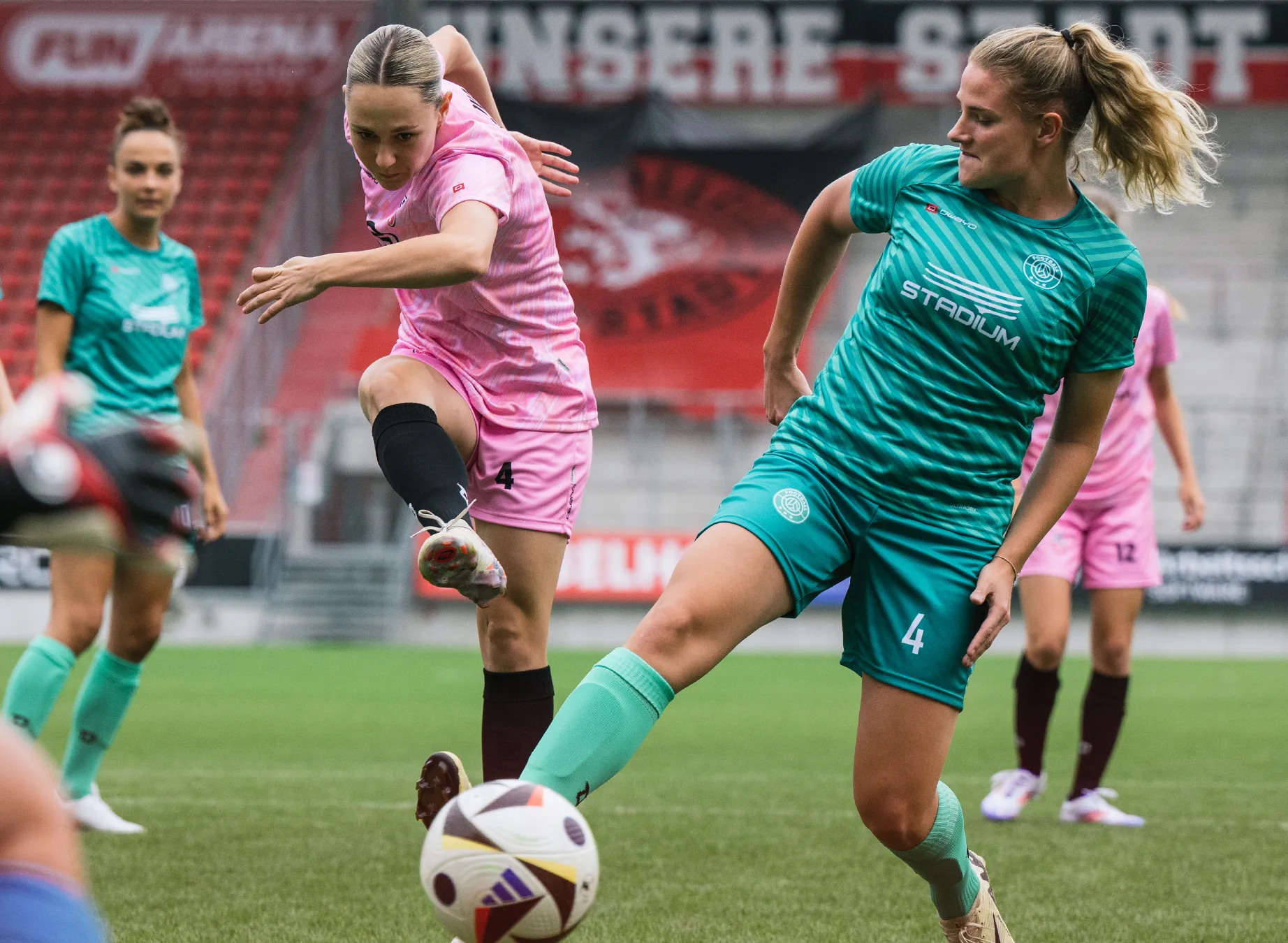 Due calciatrici in maglie vivaci, una in rosa e l'altra in verde, stanno giocando su un campo di erba.