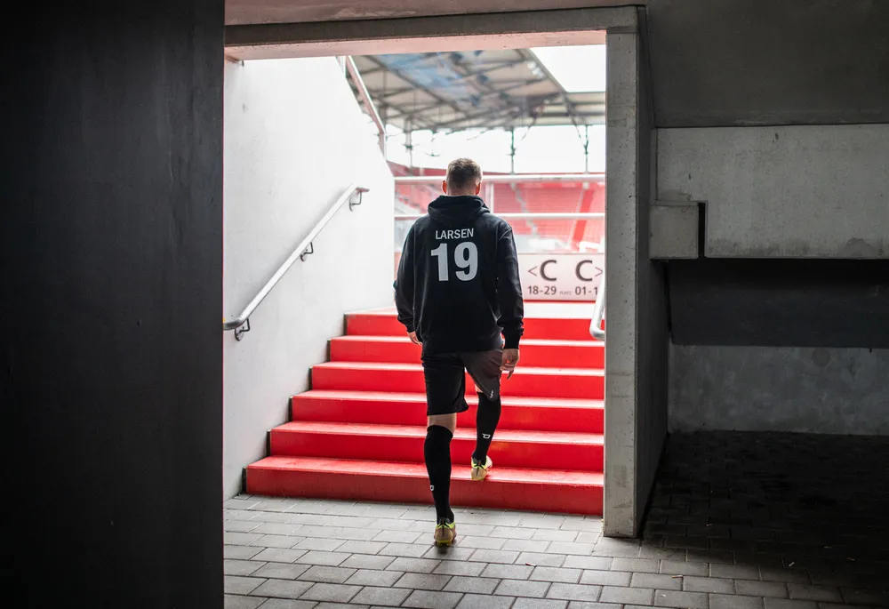 Un calciatore in un felpa nera con il numero 19 e pantaloni corti sta salendo le scale rosse di uno stadio.