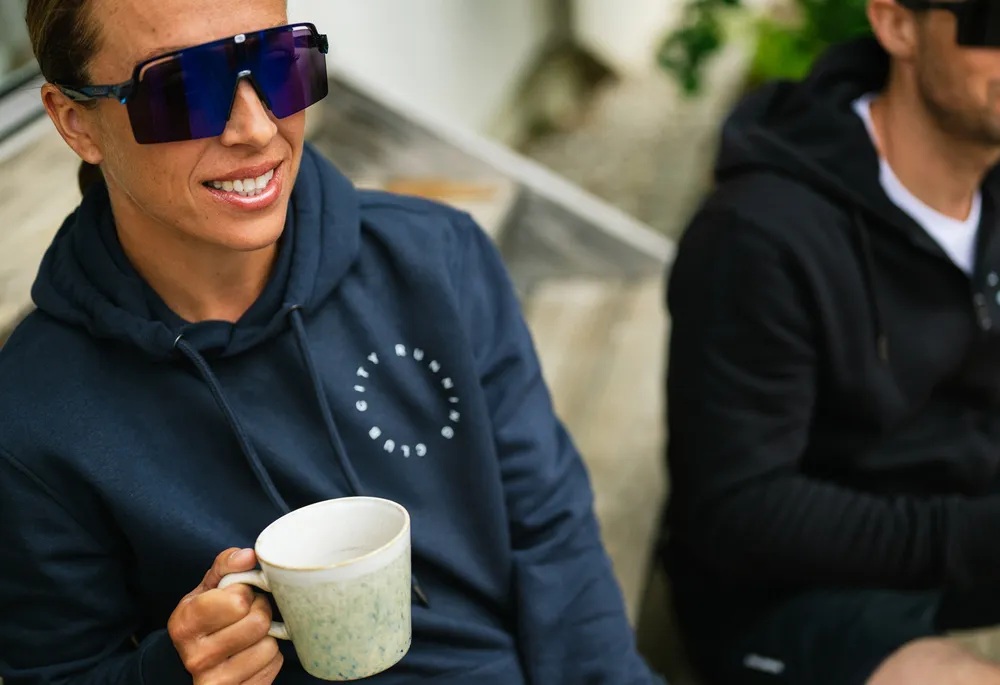 Una persona che indossa un hoodie blu con un logo tiene una tazza in mano mentre è seduta su una panchina di legno.