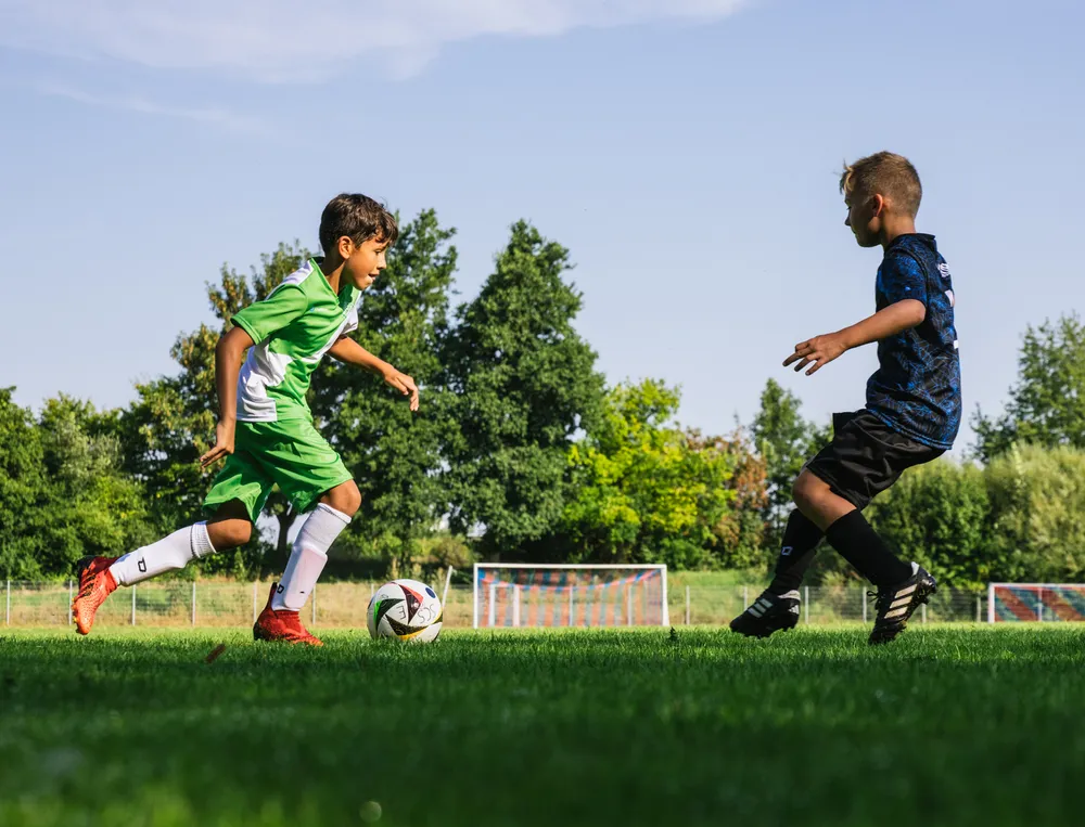 Due bambini stanno giocando a calcio su un campo verde, uno indossa una maglia verde e l'altro nera, mentre inseguono il pallone.