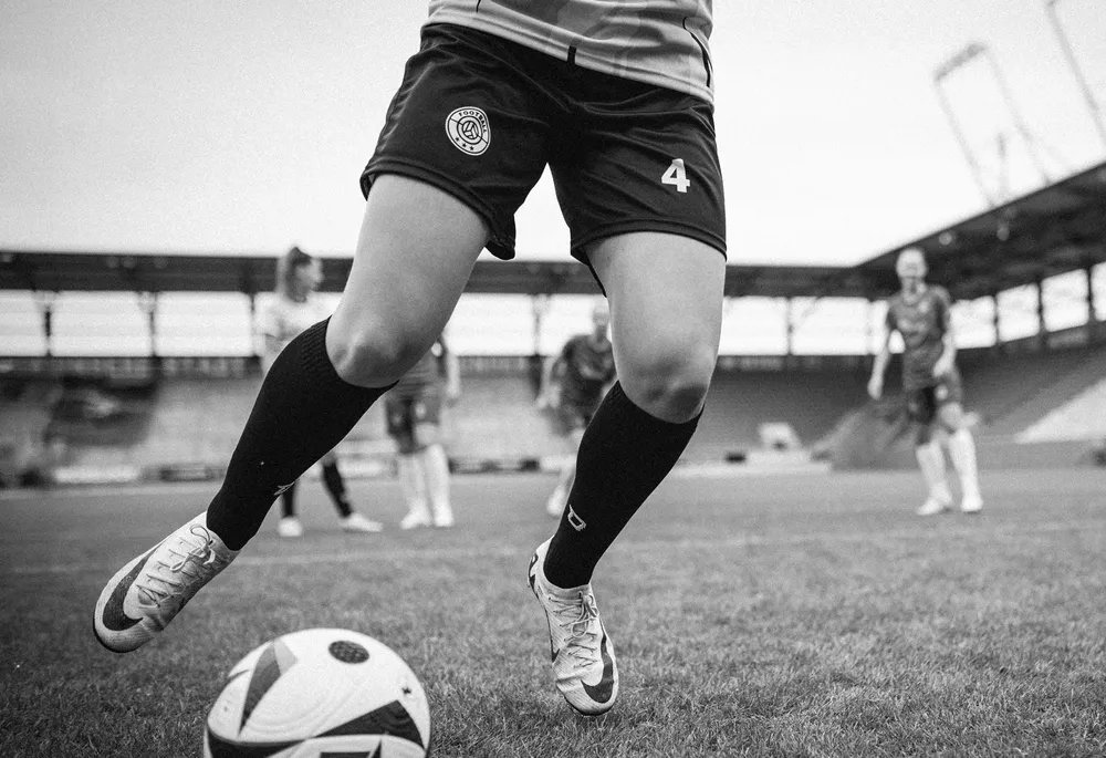 Una giocatrice con pantaloni da calcio corti e calze lunghe sta driblando un pallone sul campo.
