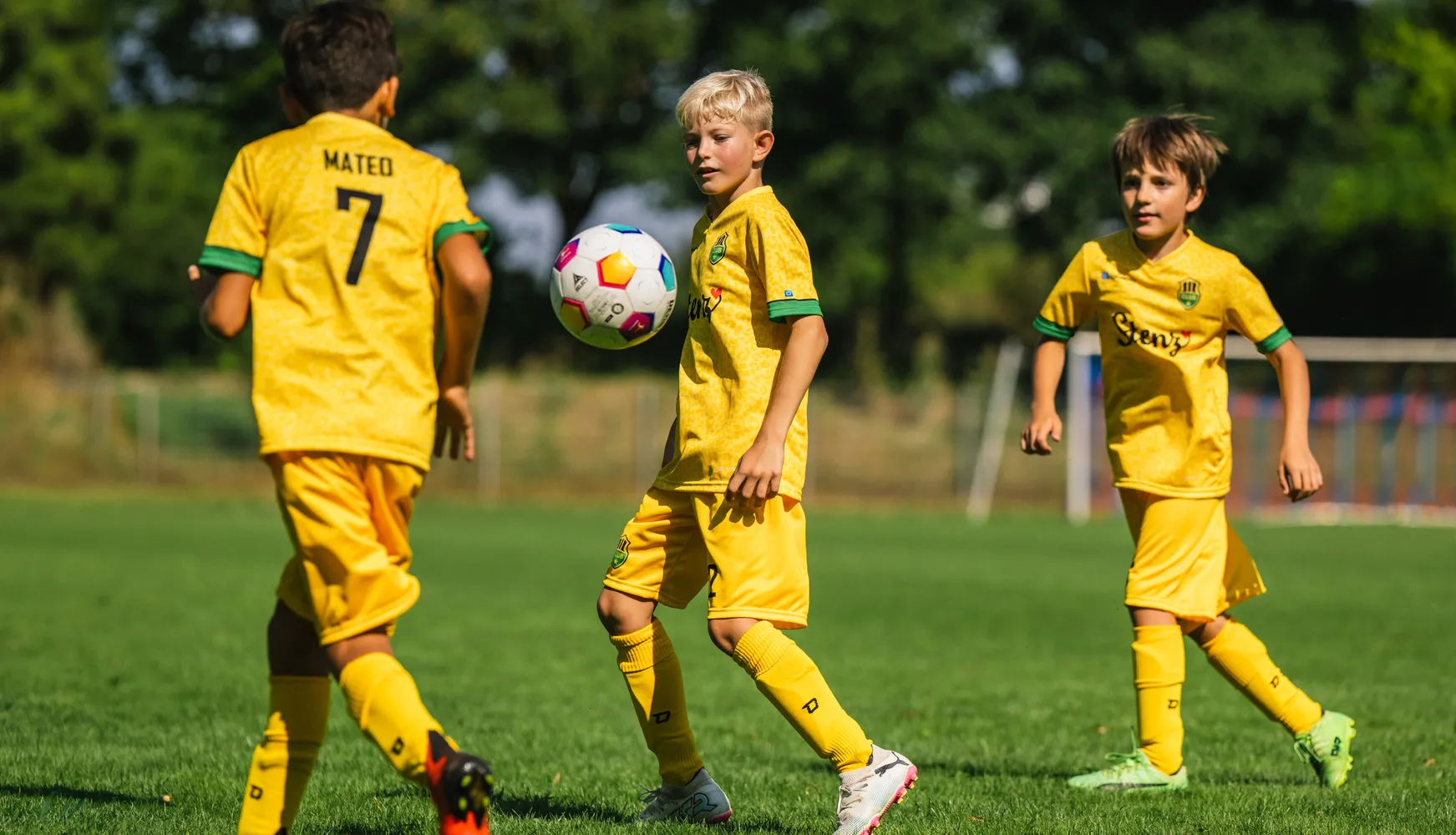 Tre bambini in magliette da calcio gialle giocano su un campo erboso con un pallone da calcio colorato.