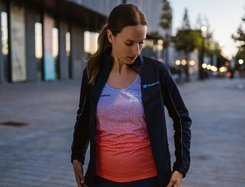 Una donna in abbigliamento sportivo, composto da una giacca leggera e una maglietta colorata, si trova in un ambiente urbano, guardando pensierosa il suo outfit.