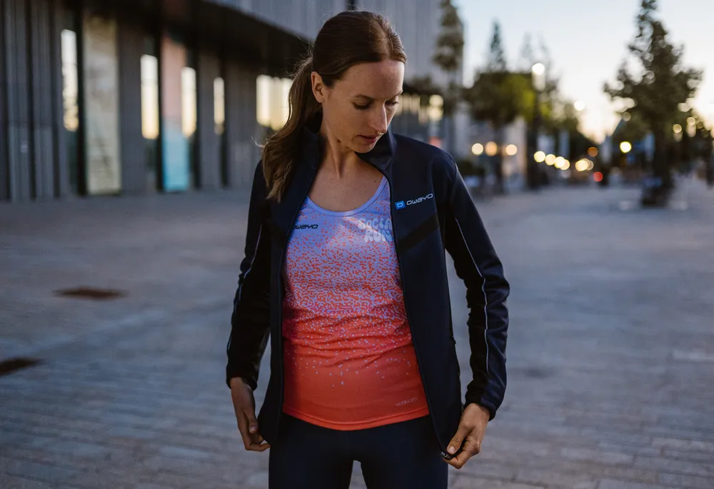 Una donna sportiva indossa una giacca blu scuro sopra una canotta senza maniche con sfumature di blu e arancione, mentre si trova in un ambiente urbano.