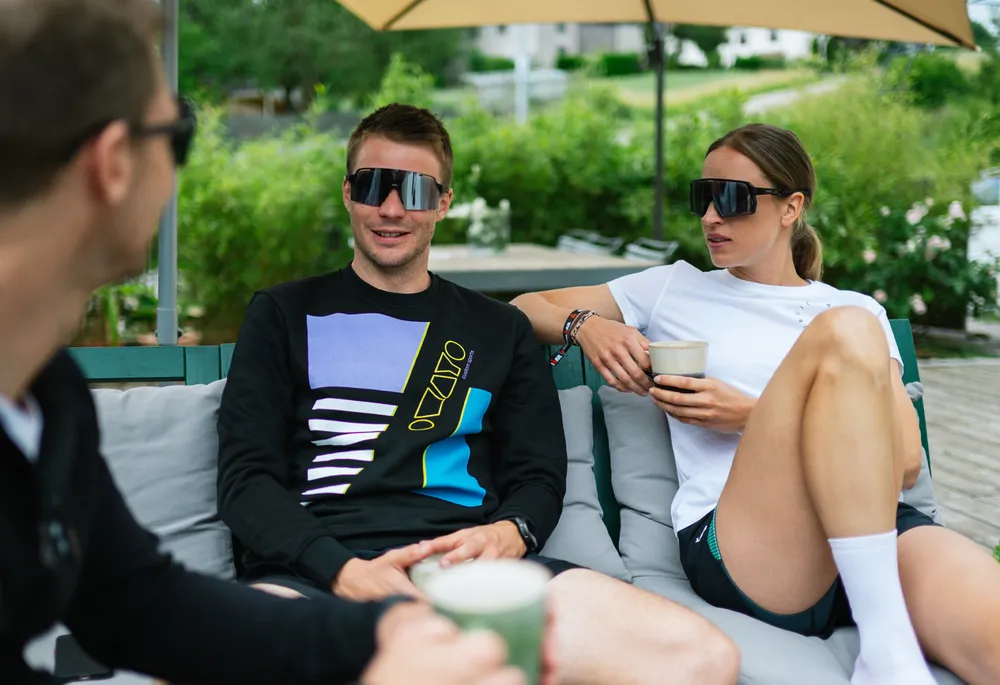 Tre persone su un divano, due vestite con abbigliamento sportivo casual e occhiali da sole, mentre si rilassano e chiacchierano tenendo delle bevande.