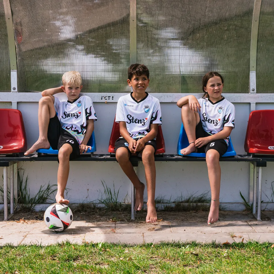 Tre bambini in magliette da calcio e pantaloni corti sono seduti su una panchina a bordo campo, con un pallone da calcio davanti a loro.