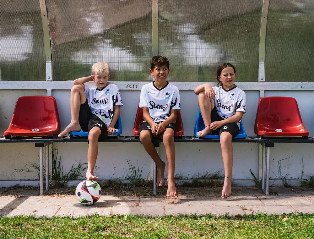 Tre bambini sono seduti su una panchina ai margini di un campo da calcio, indossando magliette da calcio personalizzate con un pallone da calcio davanti a loro.