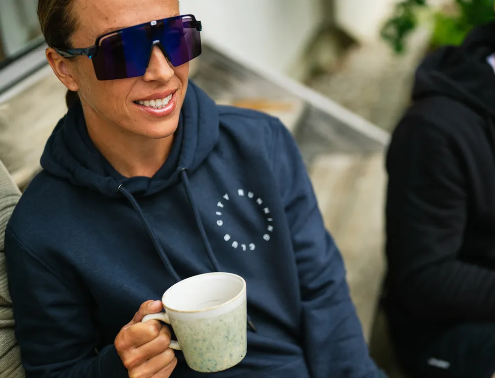Una persona in un hoodie blu navy con un logo tiene una tazza di caffè in mano e indossa grandi occhiali da sole.