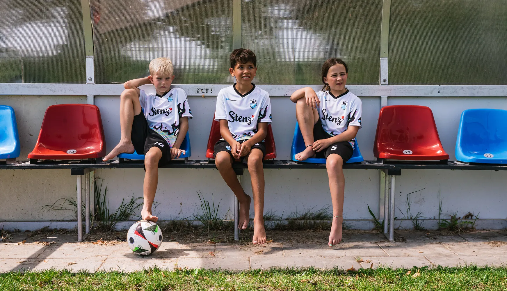 Tre bambini in magliette da calcio personalizzate sono seduti su una panchina con un pallone da calcio a terra.
