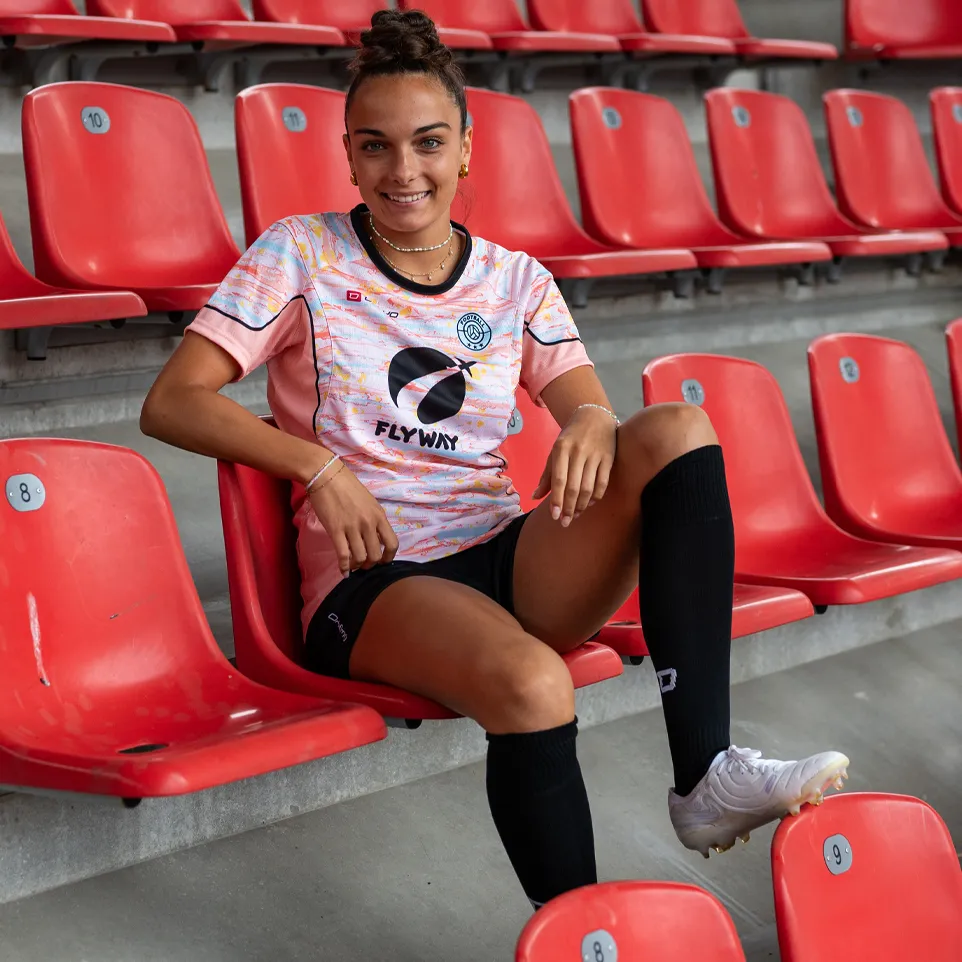 Una donna sorridente è seduta in uno stadio su una sedia rossa, indossando una maglietta da calcio colorata e calze nere.