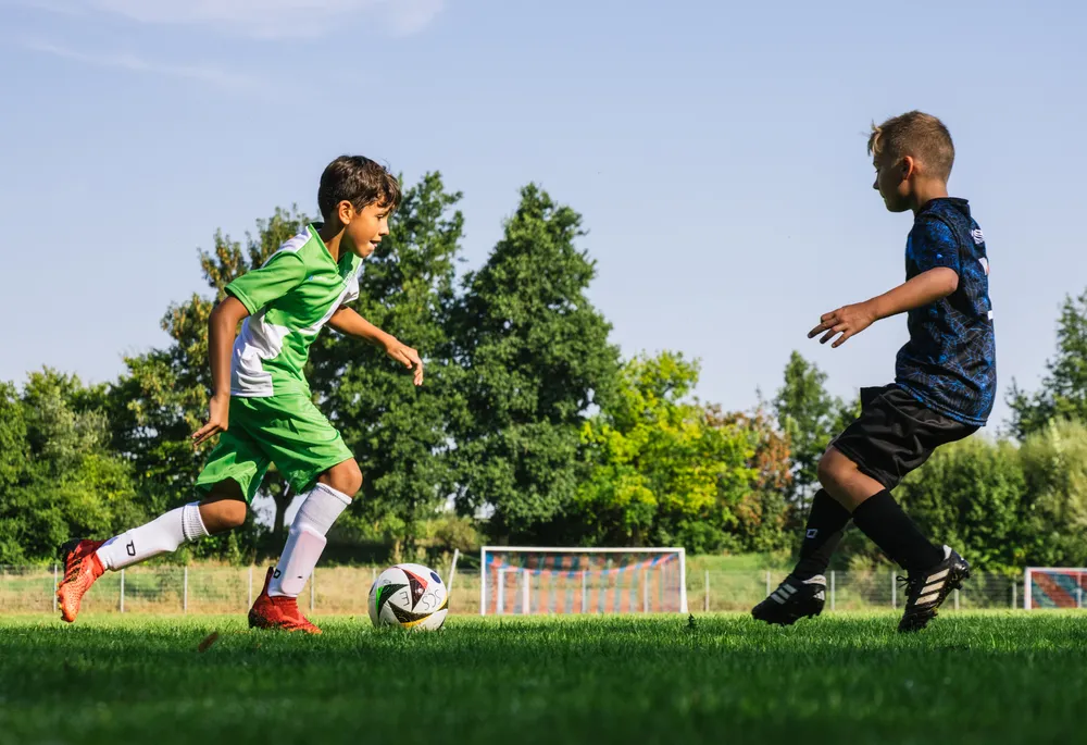 Due bambini stanno giocando a calcio su un campo verde, uno indossa una maglietta verde con accenti bianchi e l'altro una maglietta blu, mentre inseguono un pallone.