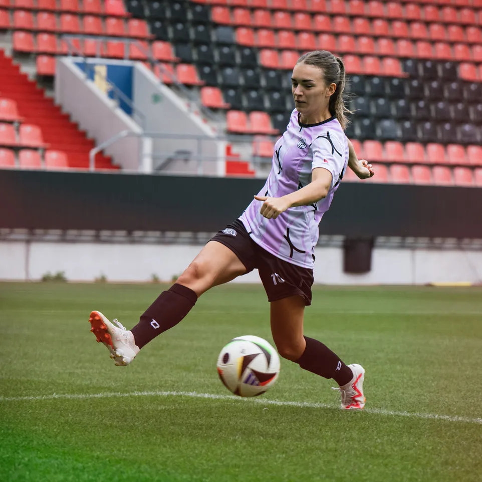 Una giocatrice calcia un pallone da calcio su un campo di stadio, indossando una maglietta viola e pantaloni neri.