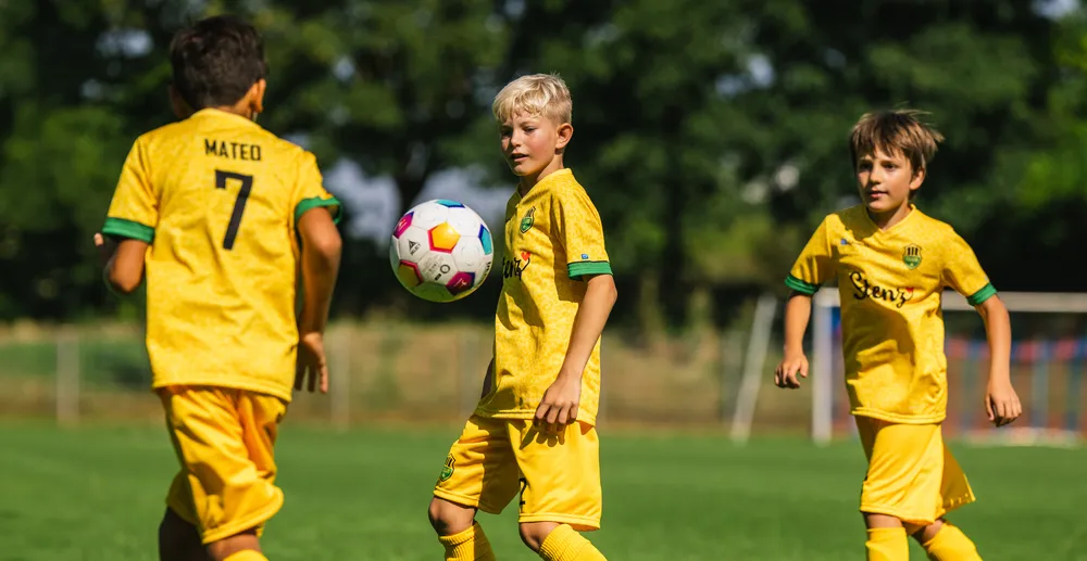 Tre bambini in magliette da calcio gialle giocano su un campo verde, mentre uno di loro controlla un pallone da calcio colorato.