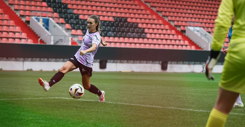 Una giocatrice in una maglietta da calcio viola e bianca calcia il pallone su un campo di stadio, con un portiere in abbigliamento giallo sullo sfondo.