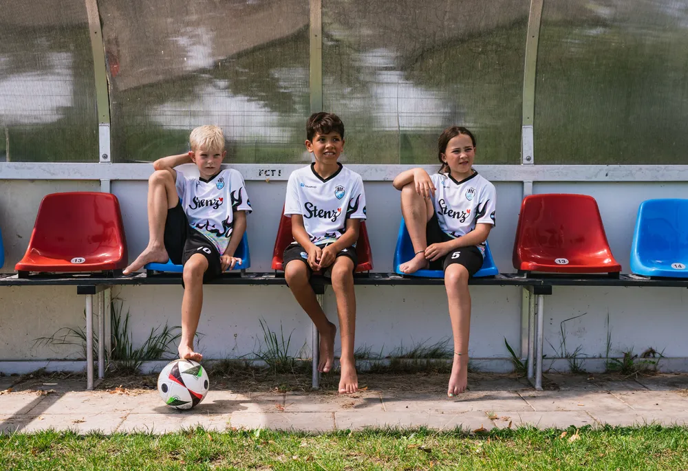 Tre bambini in magliette da calcio sportive sono seduti su una panchina accanto a un pallone da calcio.