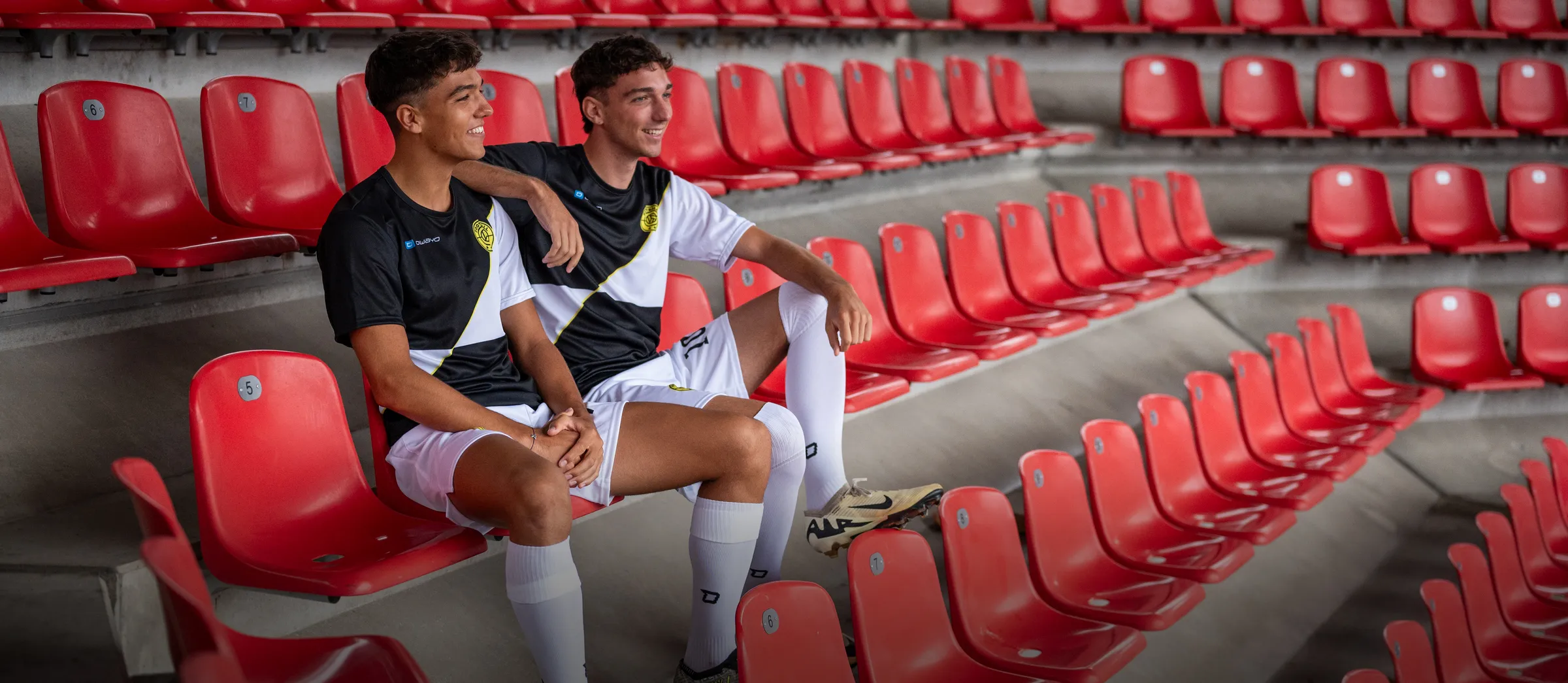 Due giovani uomini in magliette da calcio sportive sono seduti in modo informale su sedili rossi dello stadio.