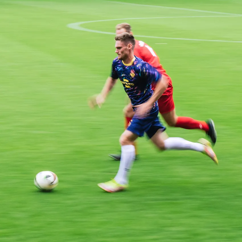 Un calciatore in una maglietta colorata corre sul campo verde mentre un altro giocatore lo insegue.