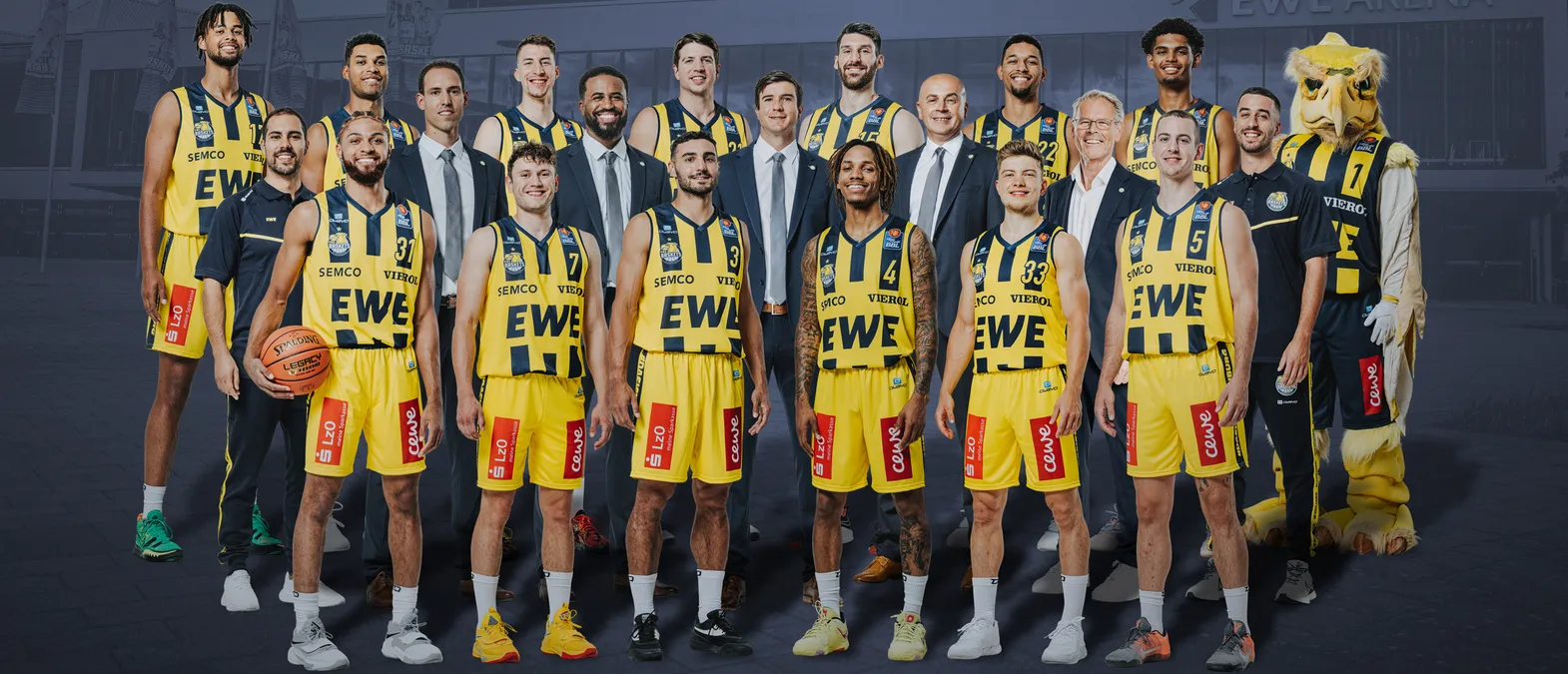Una foto di gruppo di una squadra di pallacanestro con maglie gialle e nere con i sponsor 'EWE' e 'SEMC', una mascotte e un pallone da basket, davanti a un edificio.