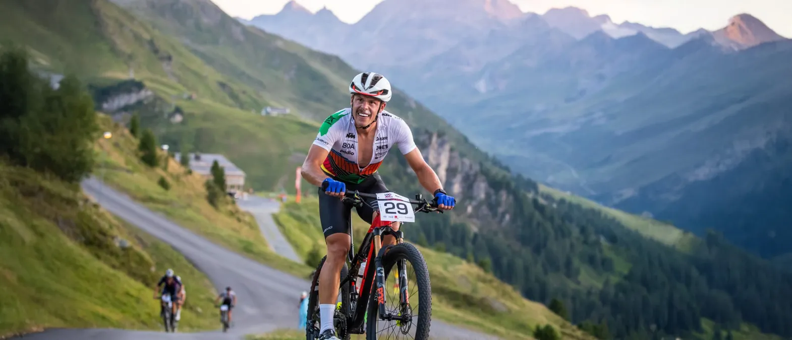 Un ciclista in una maglia bianca, verde e arancione con il numero 29 pedala su una strada di montagna in un paesaggio montuoso.