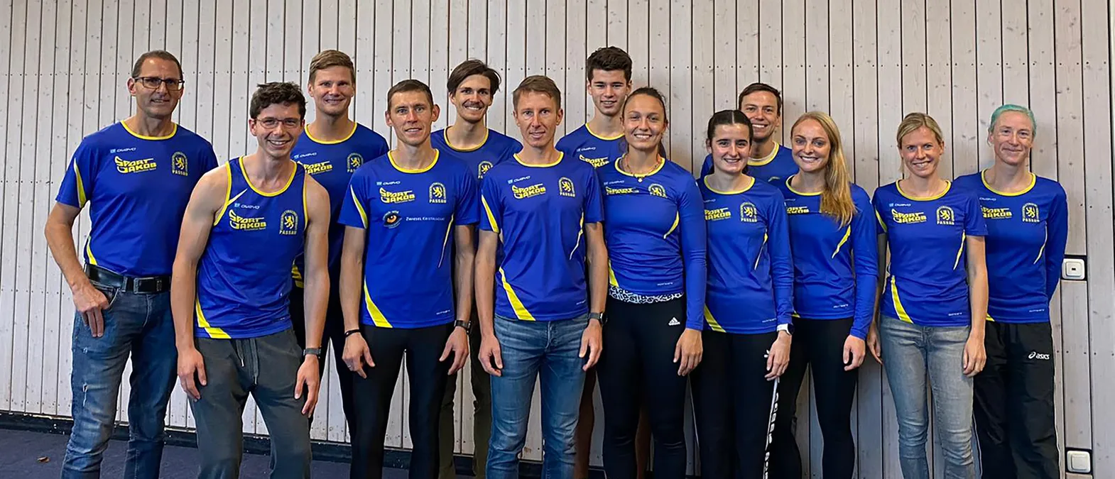 Un gruppo di uomini e donne in abbigliamento sportivo blu e giallo con il logo 'Sport Jakob Passau' posano davanti a una parete in legno chiaro.