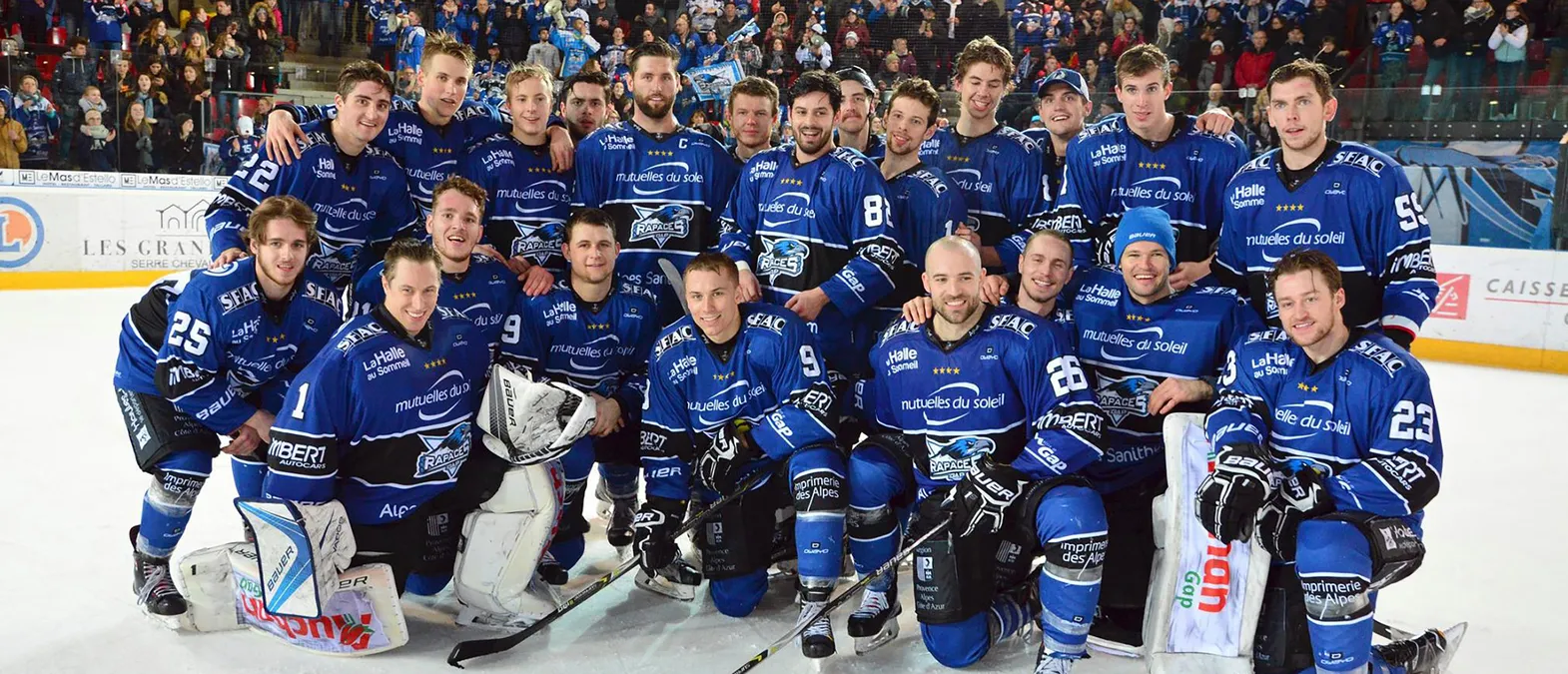 Una squadra di hockey su ghiaccio con maglie blu e il logo dei Rapaces de Gap posa sul ghiaccio davanti a un pubblico.