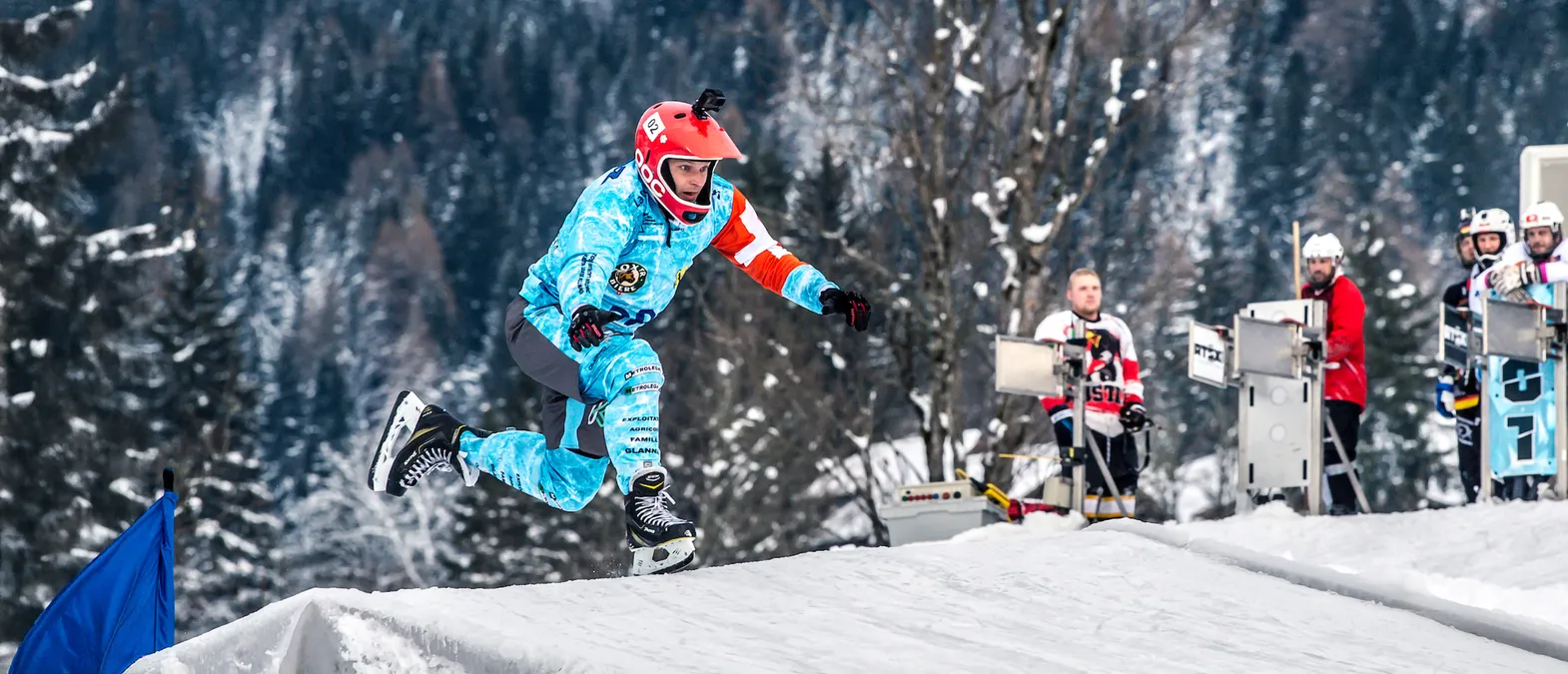 Un giocatore di hockey su ghiaccio con una maglia blu e bianca personalizzata con loghi di sponsor e un casco rosso salta su una rampa di neve su una pista di ghiaccio, con altri giocatori e alberi sullo sfondo.