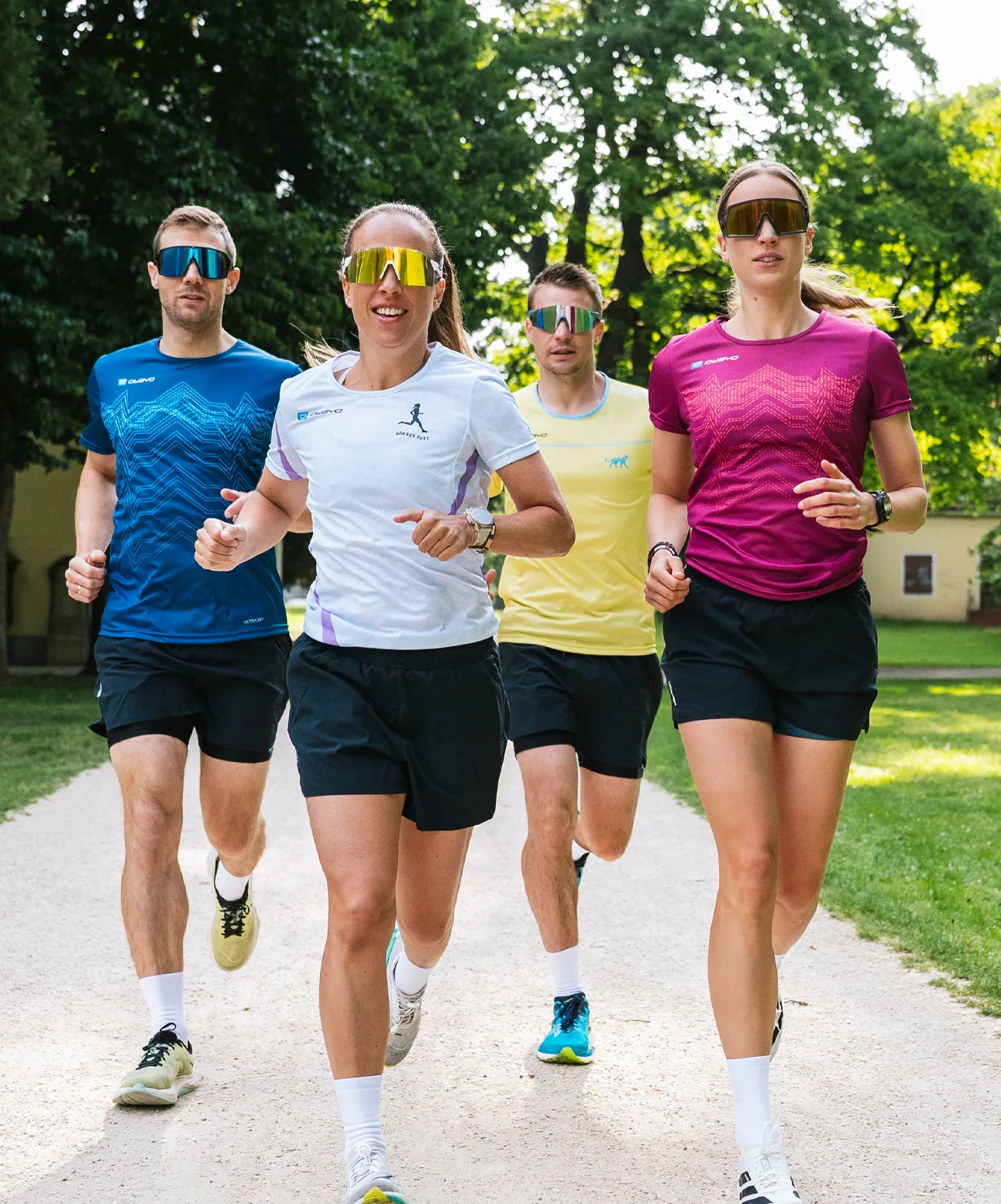 Quattro persone atletiche corrono su un sentiero stretto in un parco, indossando magliette colorate da corsa e occhiali da sole.