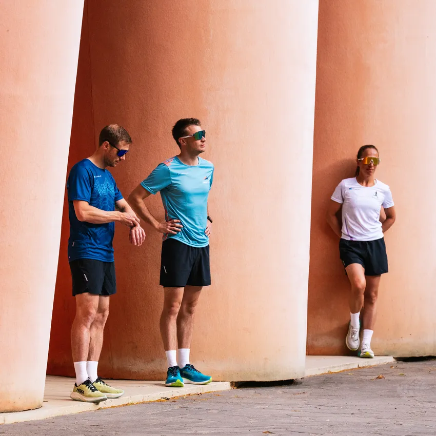 Tre atleti in abbigliamento sportivo e occhiali da sole si appoggiano in modo informale a colonne rosa.